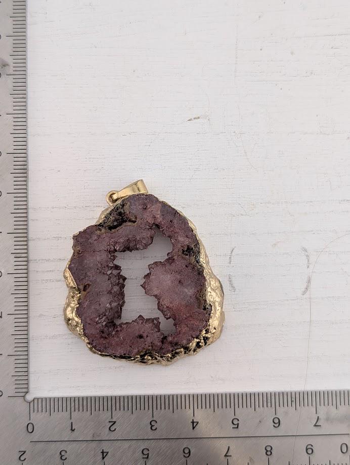 A close-up image of a purple gemstone pendant with a gold-colored frame, placed on a white surface next to a ruler for scale.