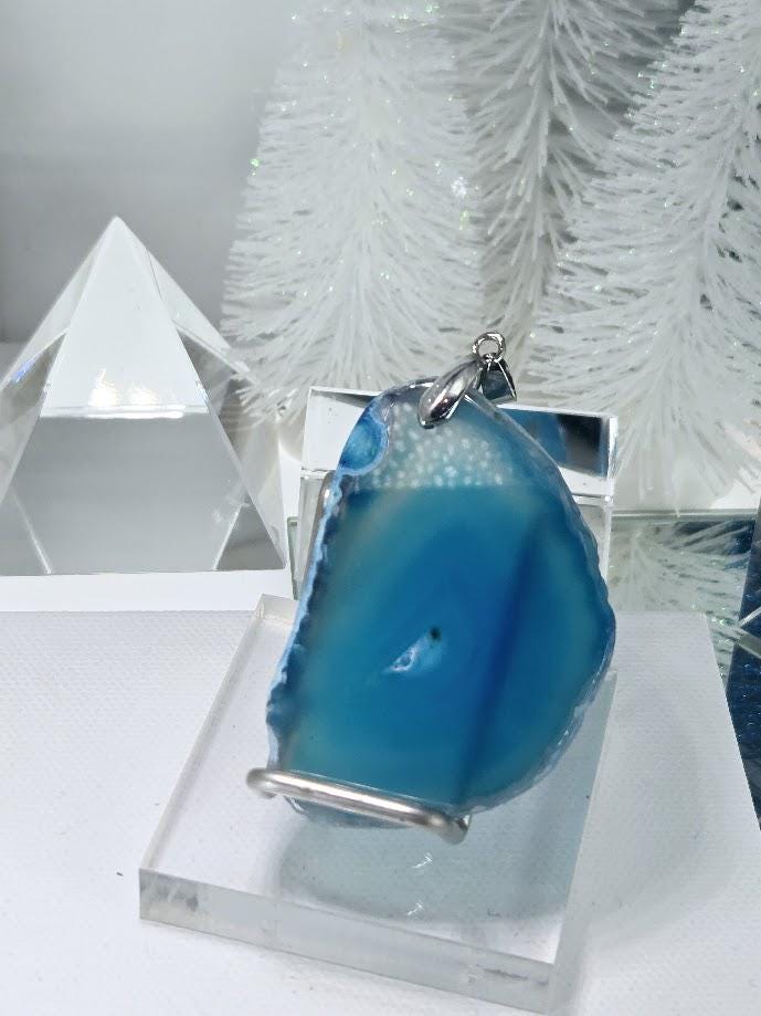 a blue agate stone on a clear stand, with a silver pendant hanging from it. The background includes a white crystal and a small white tree.