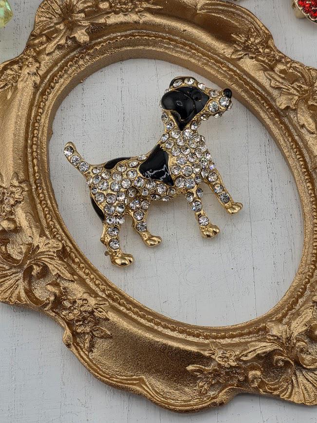 A decorative gold-framed brooch in the shape of a dog is displayed on a white surface.