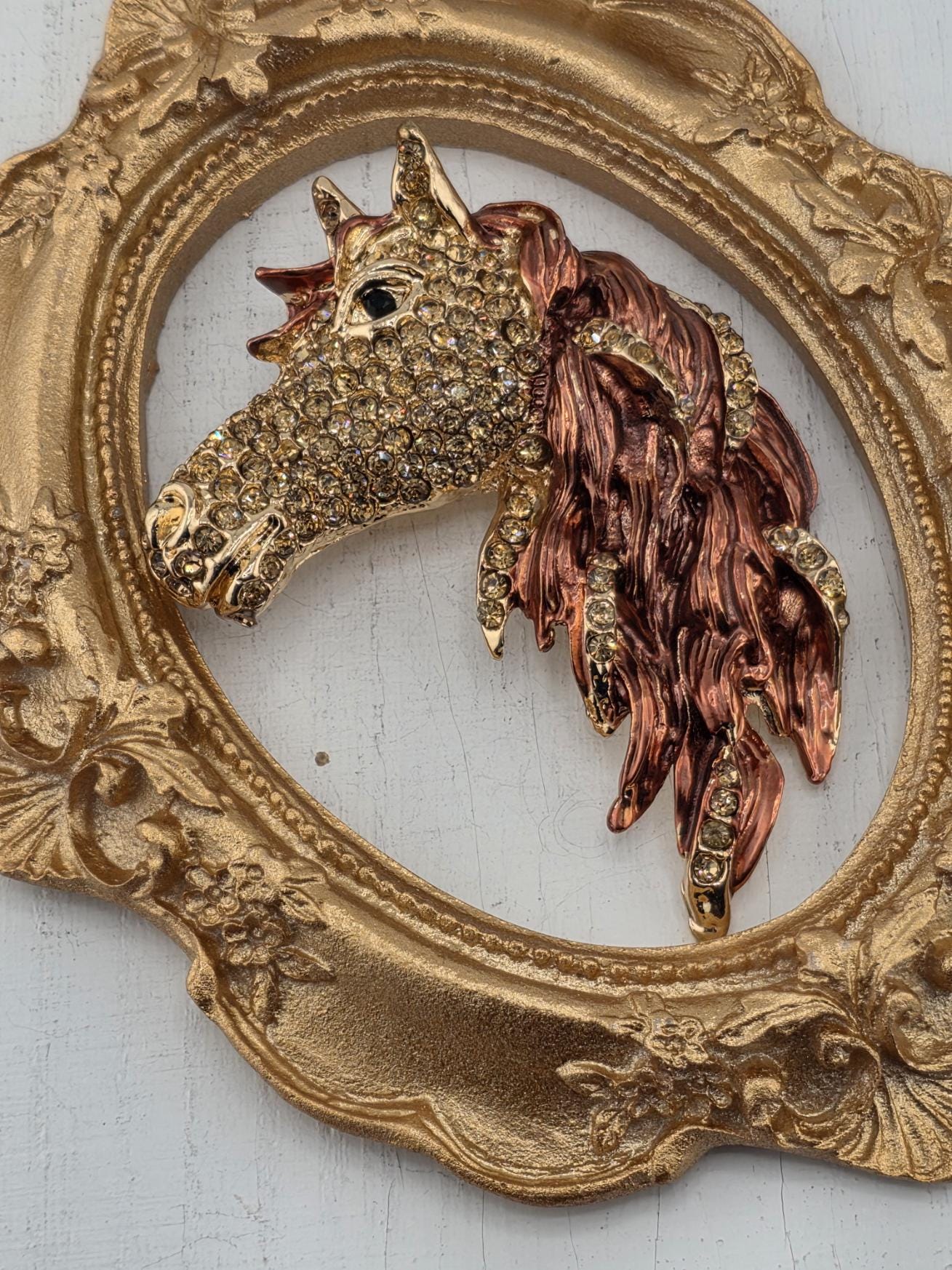 a decorative gold frame with a detailed design, including a central ornamental piece resembling a horse's head adorned with sparkling rhinestones.
