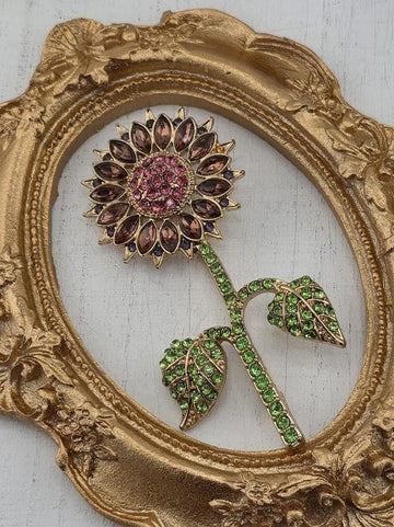 A decorative gold-colored oval frame with a large, ornate floral brooch featuring a pink and purple flower with green leaves.