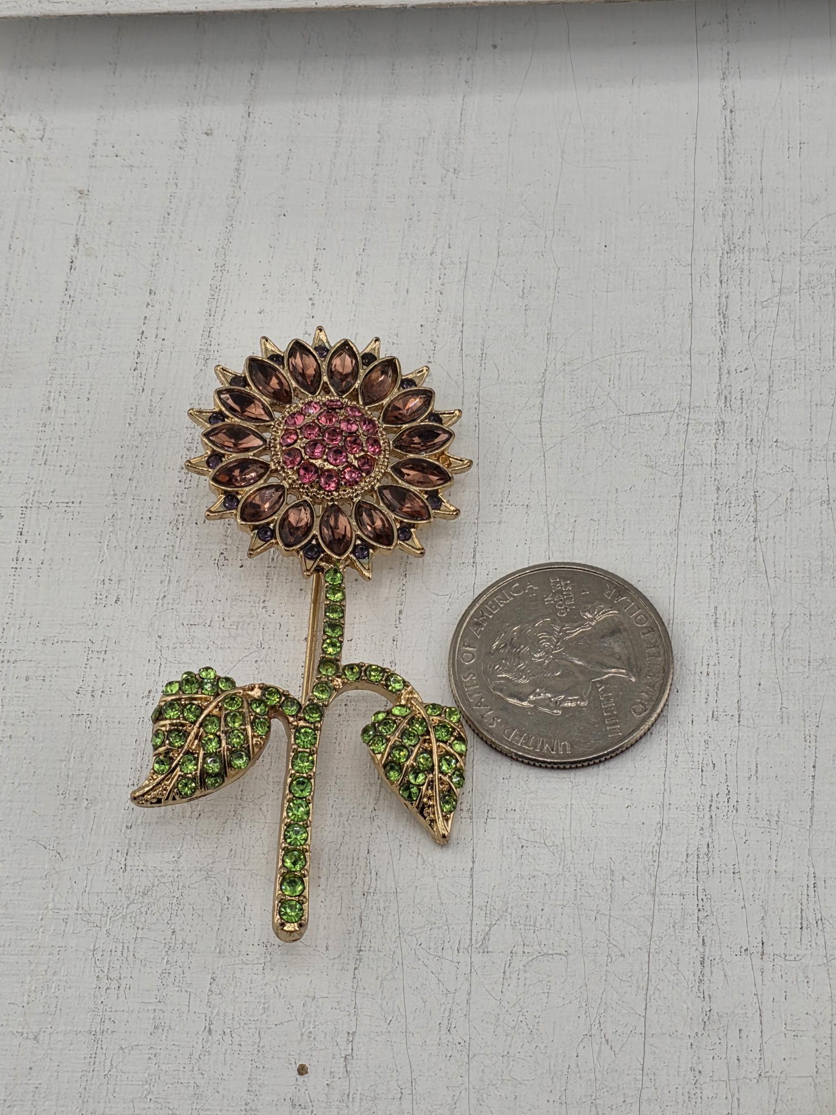 A decorative brooch with a flower-like design and green gemstones is placed next to a United States quarter coin on a white wooden surface.
