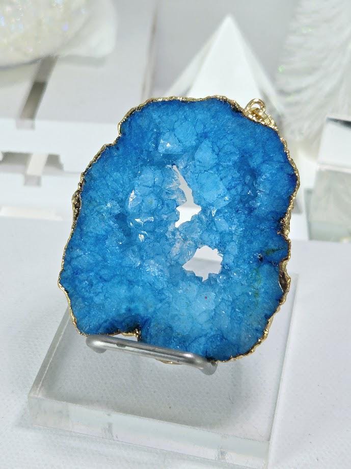 A close-up image of a blue geode pendant with a gold-colored metal frame, placed on a white surface.