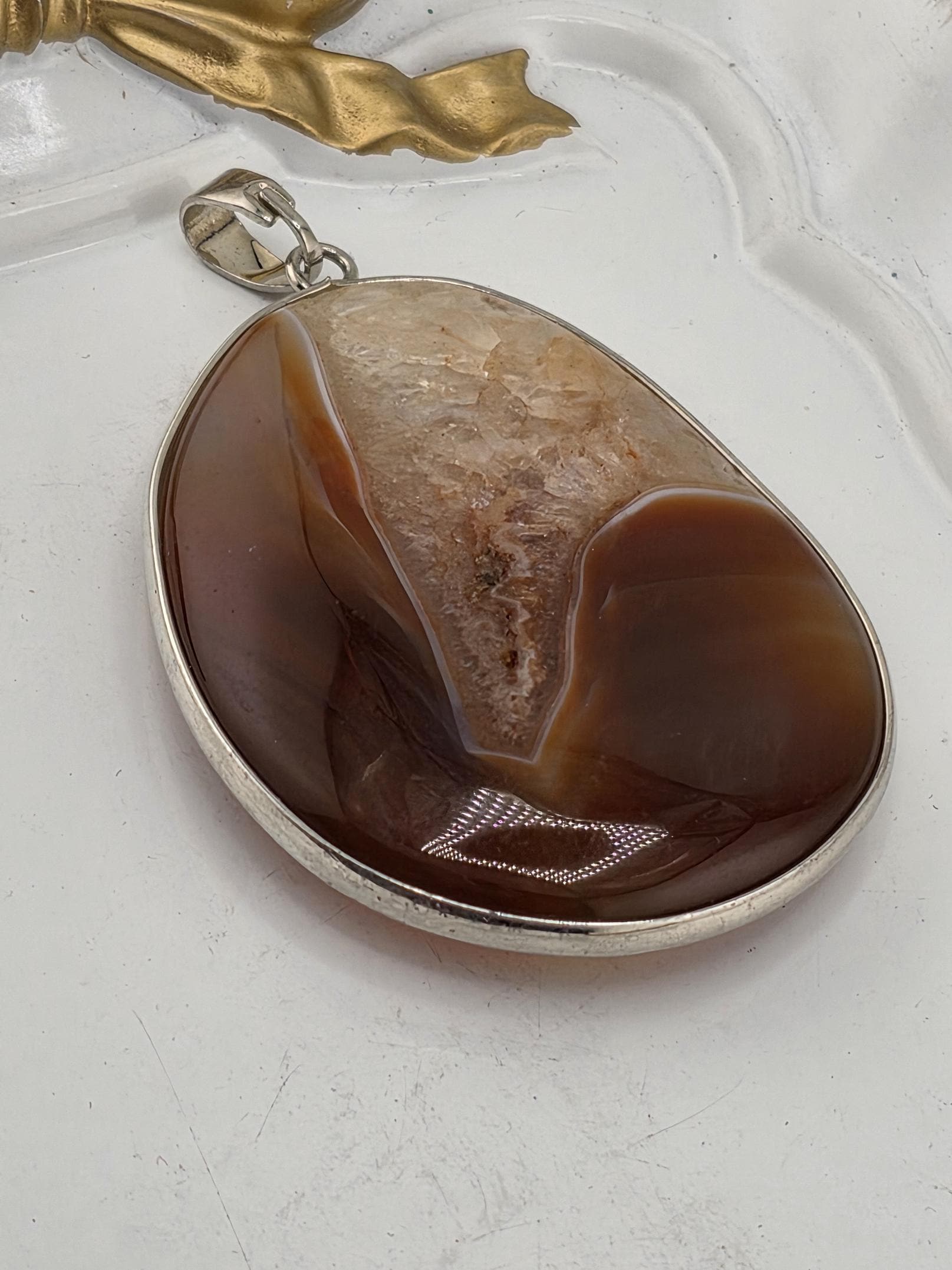 A large oval-shaped stone with a brownish-brown hue, possibly a piece of amber or a similar type of rock, is displayed on a white surface. The stone is encased in a silver frame, and there is a small gold-colored leaf or branch attached to the frame.