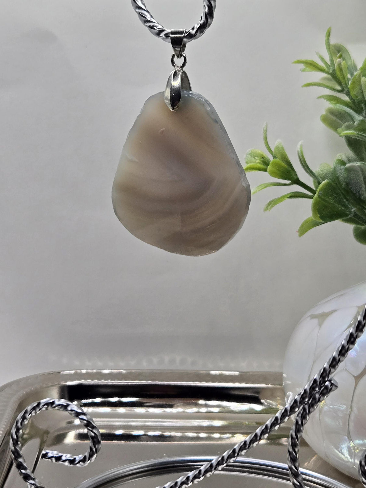 a close-up view of a pendant with a unique, striped stone shape hanging from a chain. The stone appears to be a natural stone, possibly a piece of agate or a similar type of rock. The pendant is set against a backdrop of a white surface, with a plant visible in the background.