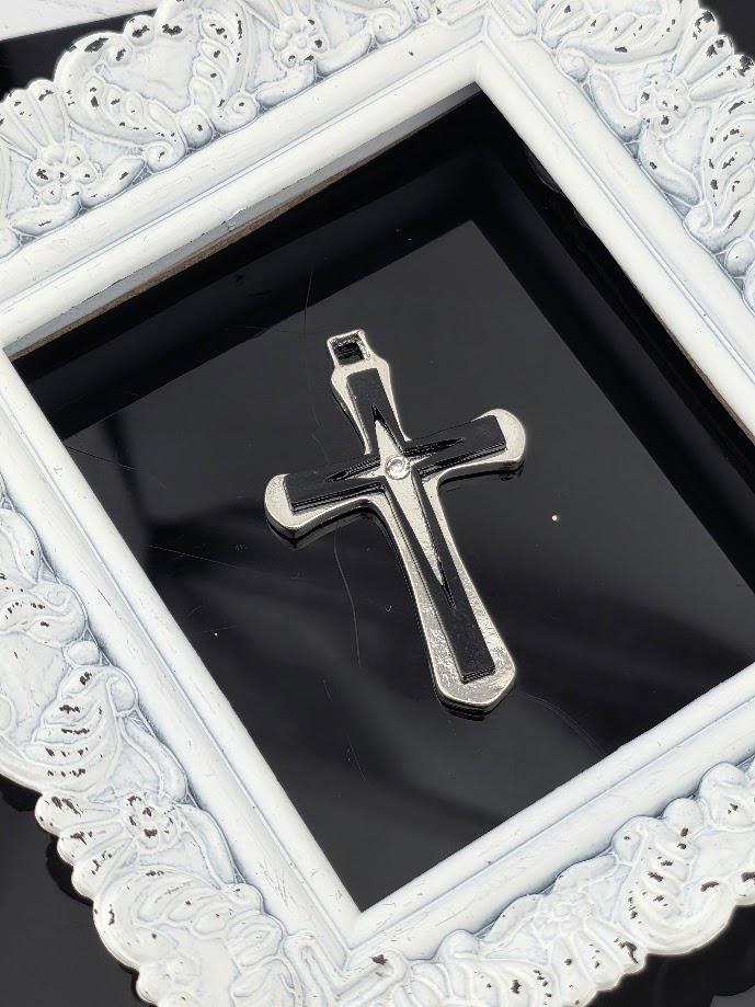 A silver cross pendant is displayed within a white ornate frame against a black background.
