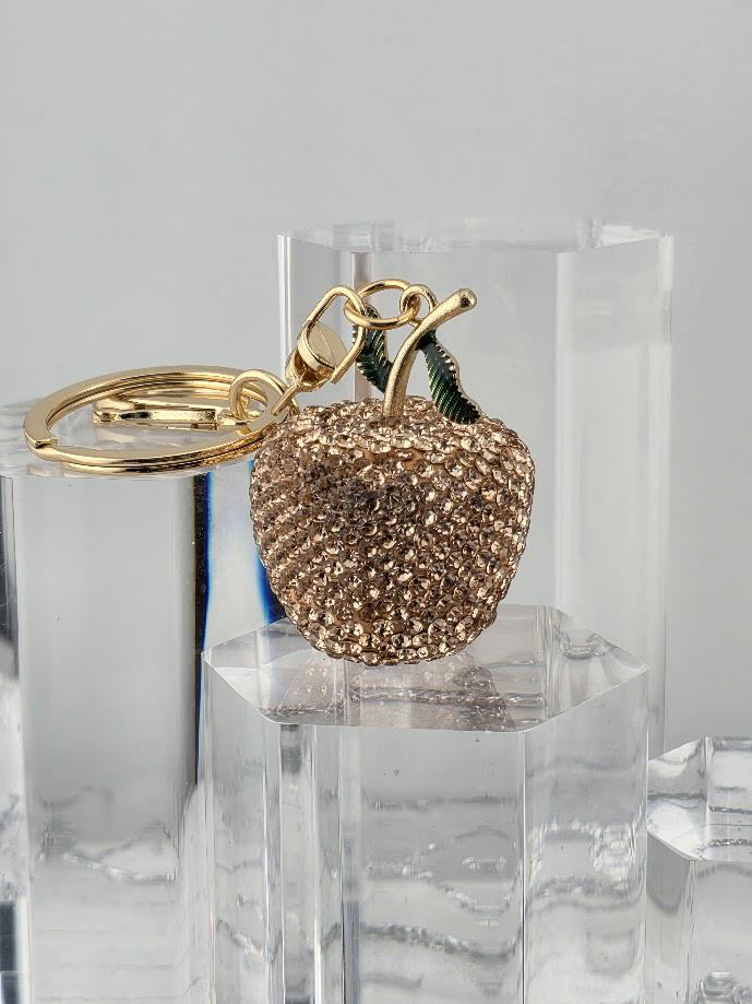 A gold-colored keychain in the shape of an apple, with a sparkling crystal-like design, is displayed in a black box.