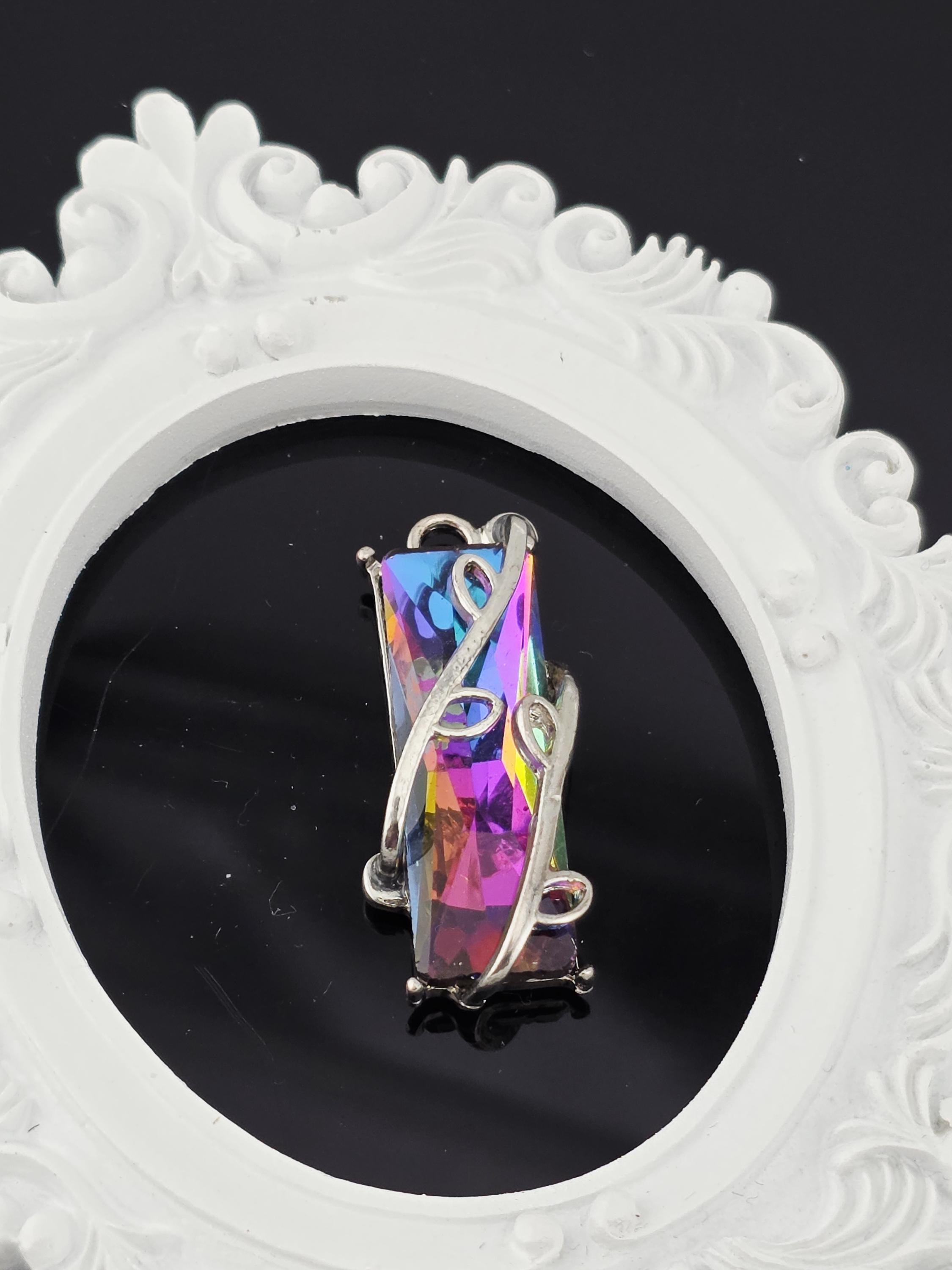 A piece of jewelry with a colorful gemstone, possibly a crystal or gemstone, is displayed against a white ornate frame.