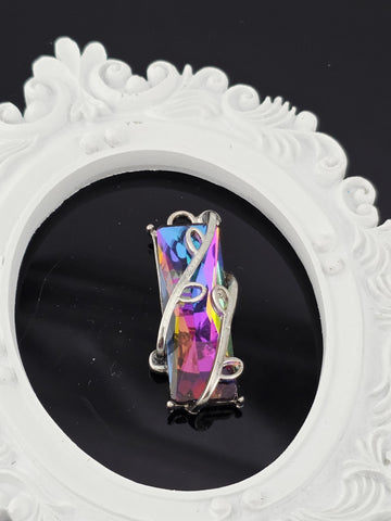 A piece of jewelry with a colorful gemstone, possibly a crystal or gemstone, is displayed against a white ornate frame.