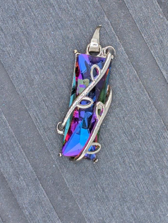 A pendant with a colorful, multicolored gemstone set in a silver metal frame, resting on a textured gray surface.