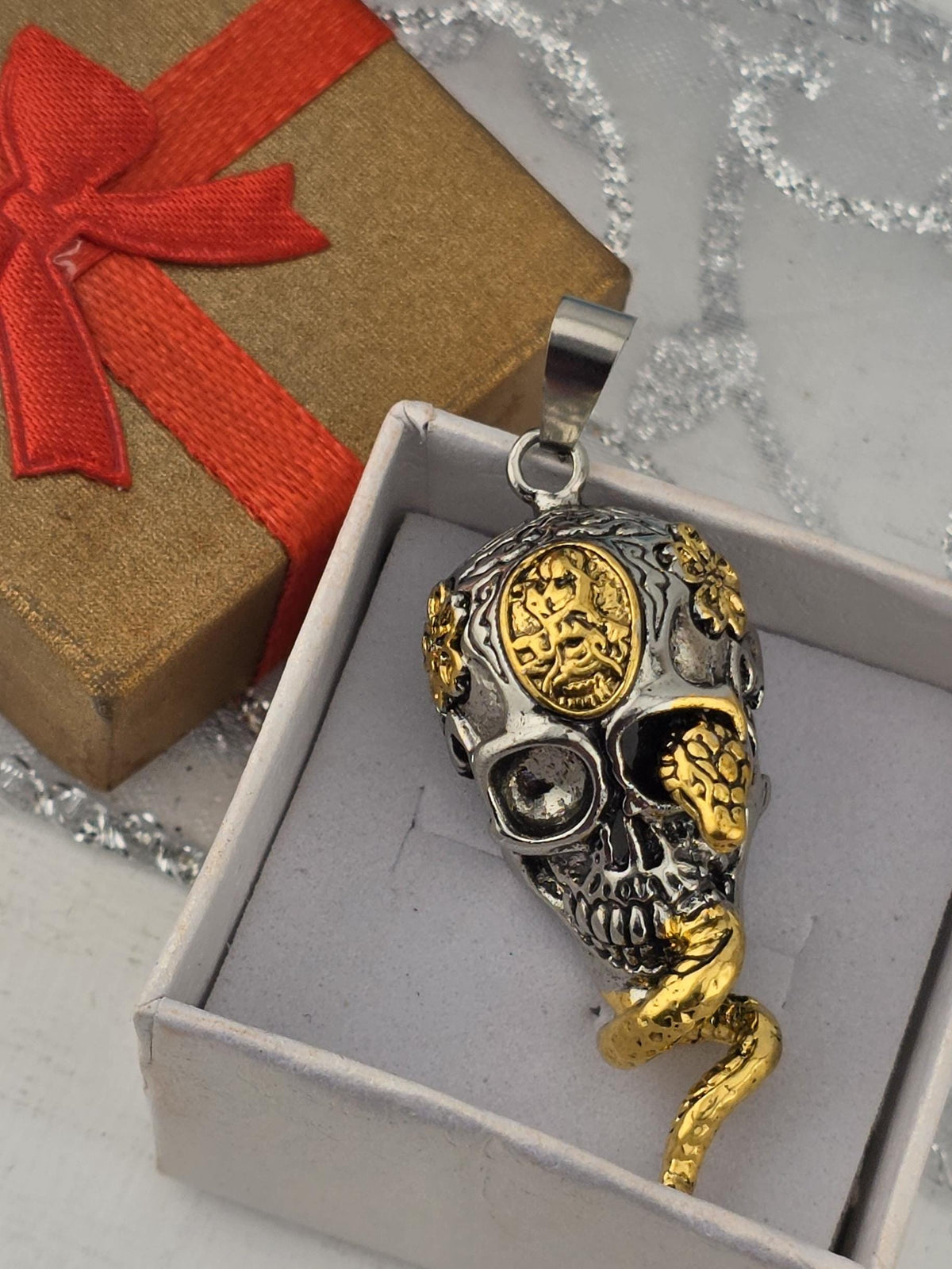 a pendant in the shape of a skull with a gold and silver design, displayed in a white box with a red ribbon.
