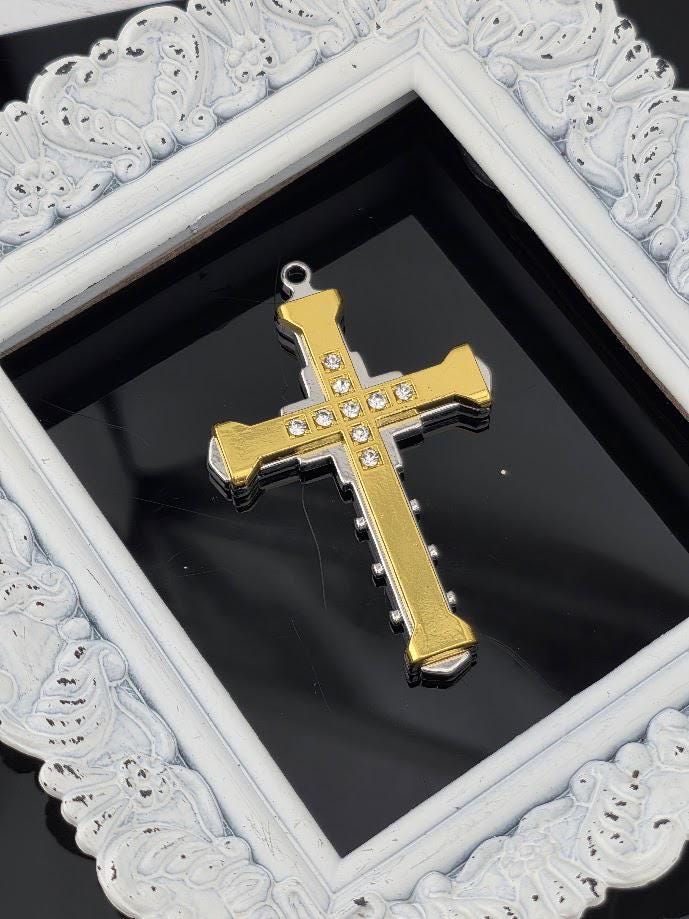 A gold and silver cross pendant with small rhinestones is displayed in a white ornate frame against a black background.
