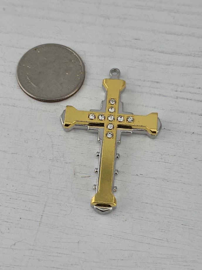 A gold and silver cross pendant with small rhinestones is displayed in a white ornate frame against a black background.