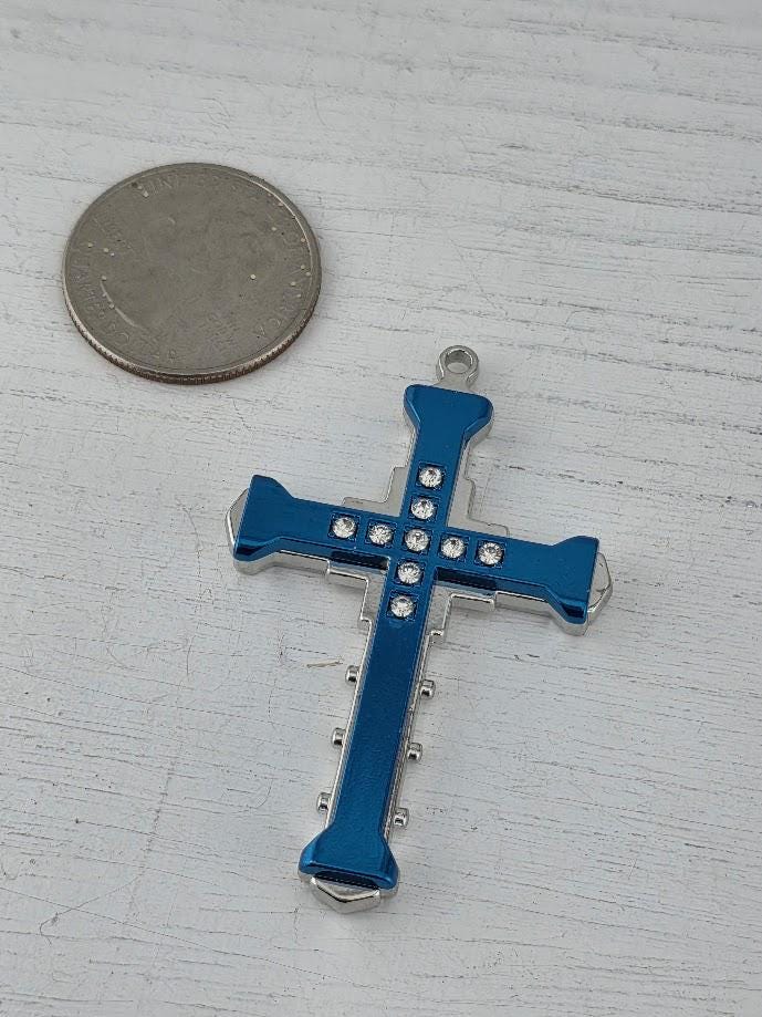 A blue cross-shaped pendant with small rhinestones or crystals is placed next to a silver coin on a white surface.