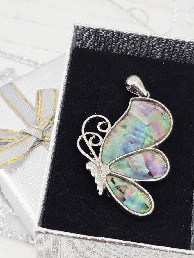 A silver butterfly pendant with colorful iridescent wings is displayed in a white jewelry box with a gold ribbon.