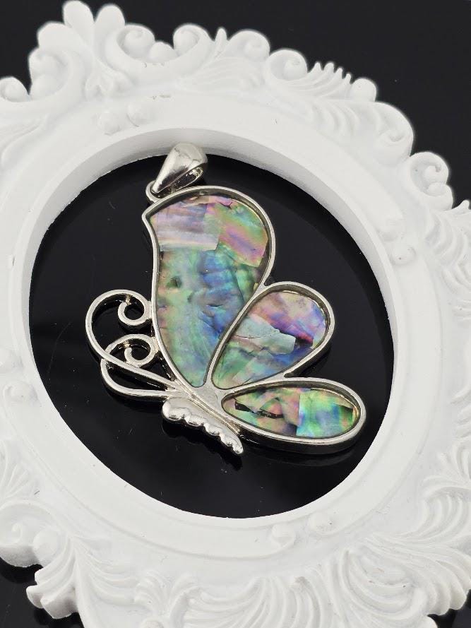 A silver-toned pendant in the shape of a butterfly, with colorful inlays, is displayed against a black background within an ornate white frame.