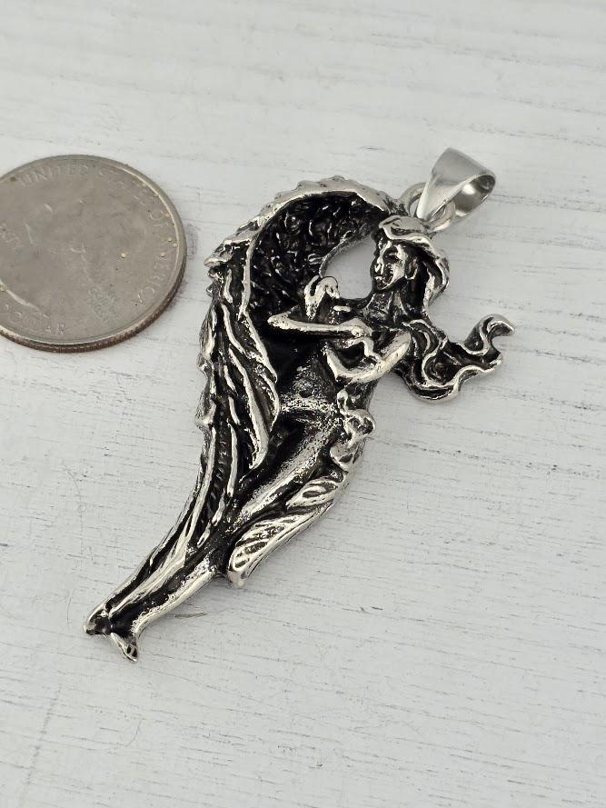 A silver-colored pendant in the shape of a winged figure, possibly an angel, is shown next to a coin on a wooden surface.
