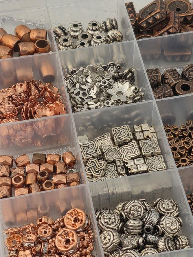 A collection of various beads and metal pieces, including copper, silver, and gold-colored beads, as well as metal beads with intricate designs, organized in multiple compartments.