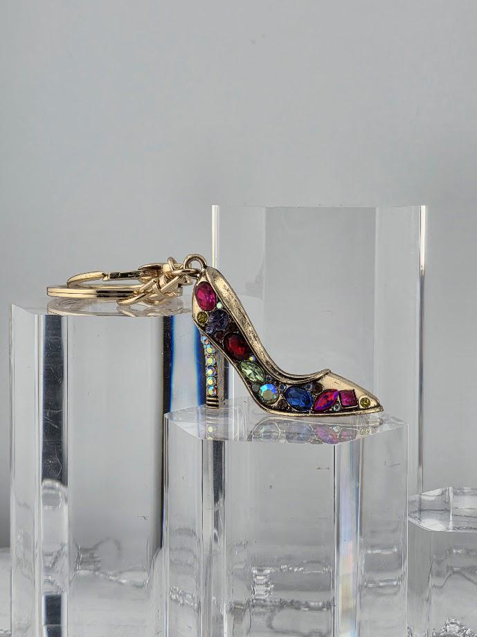 A gold-colored high-heeled shoe with colorful gemstones is displayed on a clear glass stand.