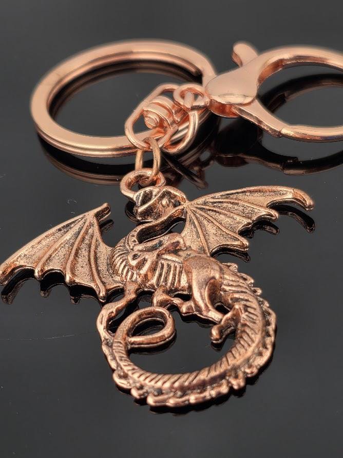 A close-up image of a copper-colored dragon keychain with wings and a tail, resting on a reflective surface.
