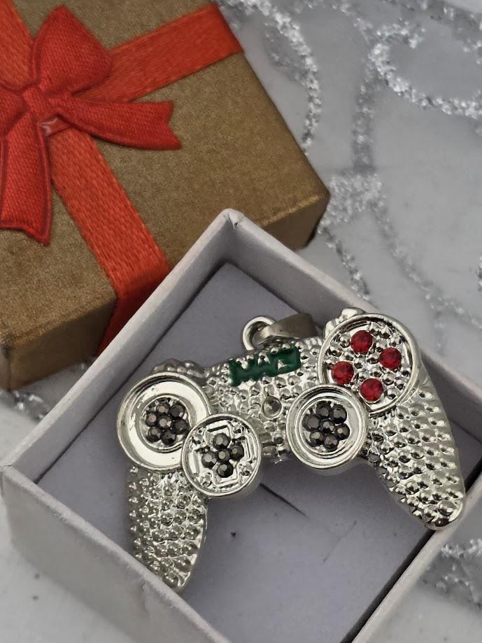A silver and green pendant in the shape of a video game controller, with red gemstones, is displayed in a white box with a red ribbon.