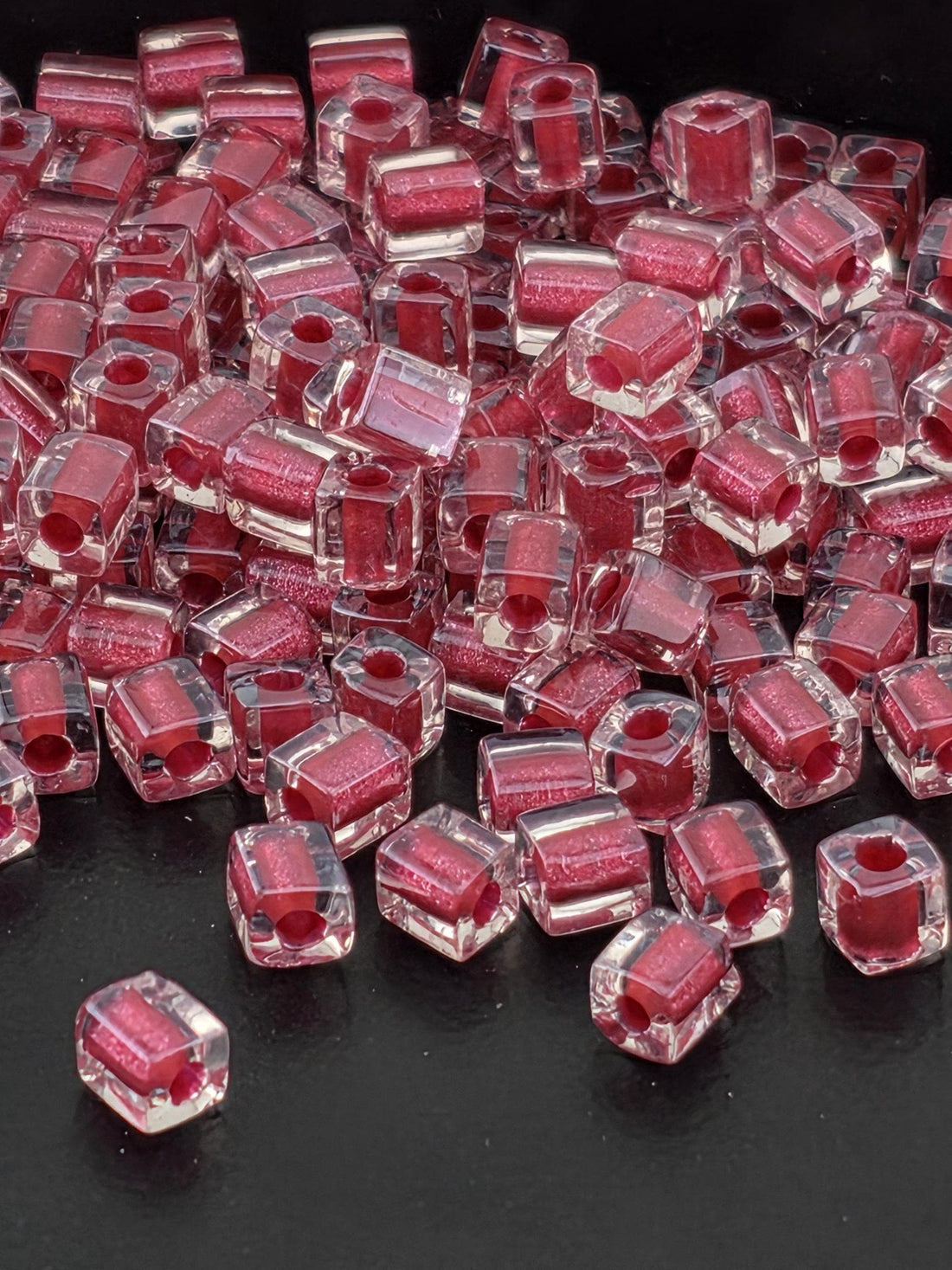 A close-up image of numerous clear, transparent beads in a vibrant red color, scattered across a dark background.