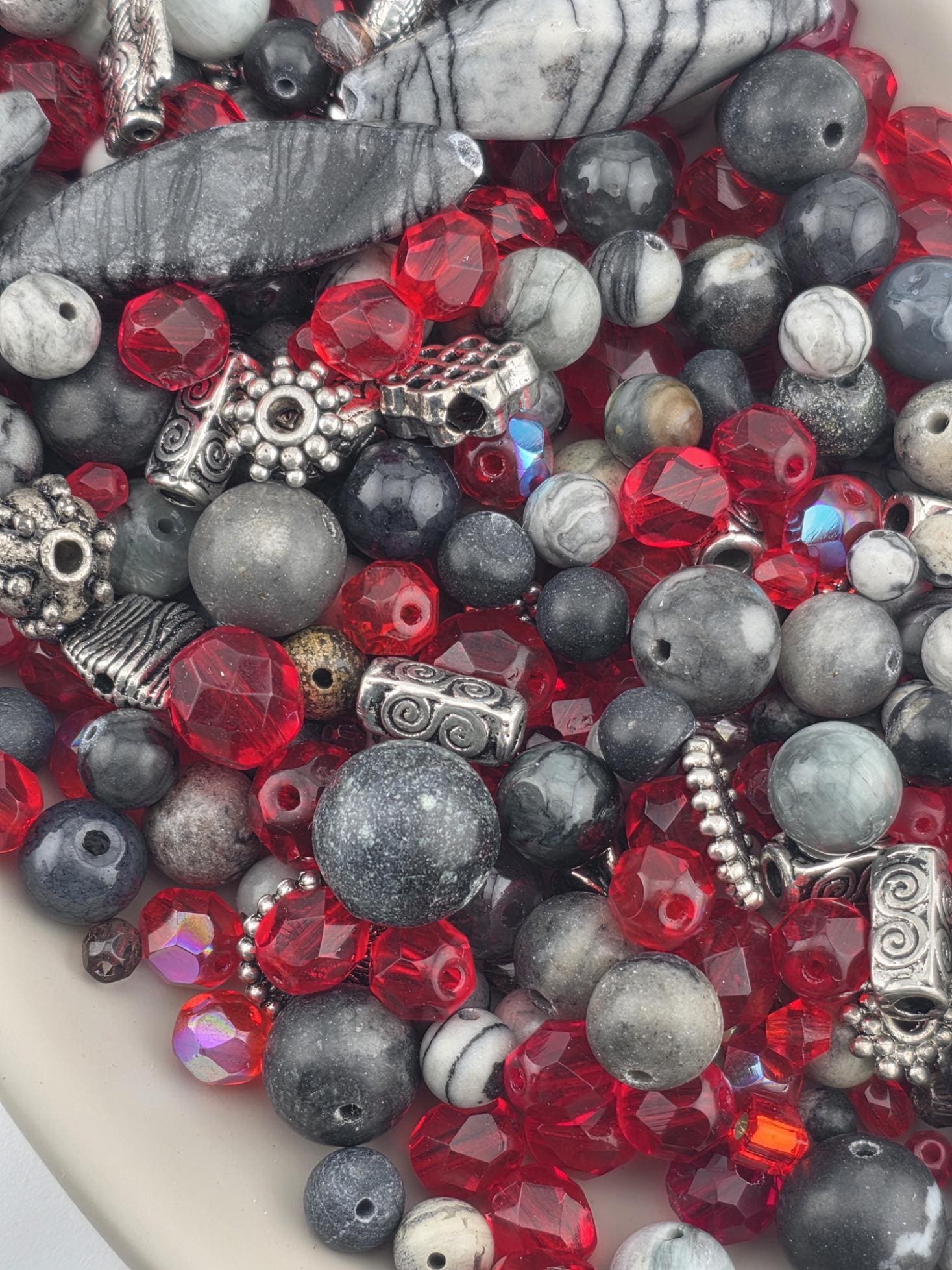 The image is a close-up of a variety of beads and beads, including red, gray, and black beads, as well as some with metallic elements. The beads are scattered and overlapping, creating a visually interesting and diverse collection.