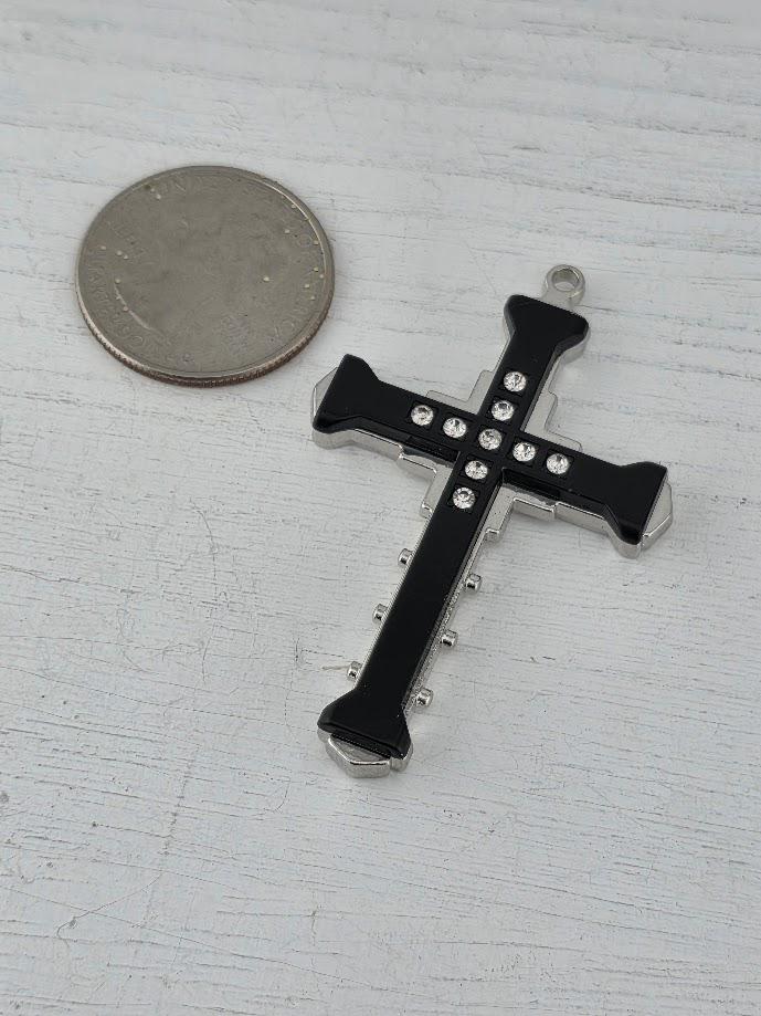 A black cross-shaped pendant with small rhinestones or crystals is placed next to a silver coin on a white wooden surface.