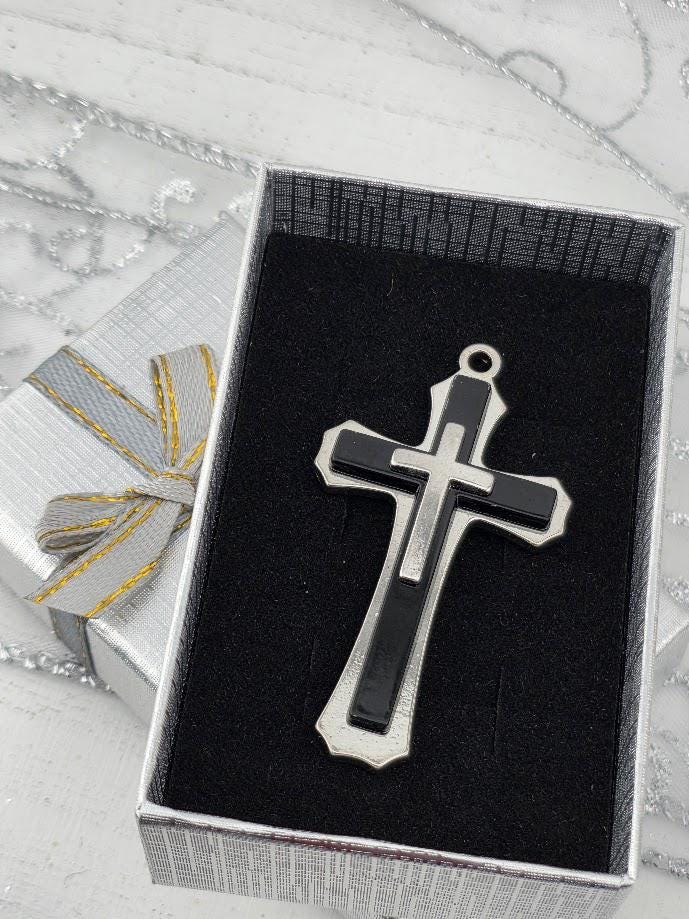 A silver and black cross pendant is displayed in a silver box with a gold ribbon bow.
