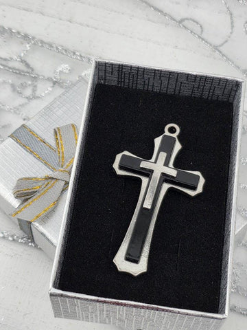 A silver and black cross pendant is displayed in a silver box with a gold ribbon bow.