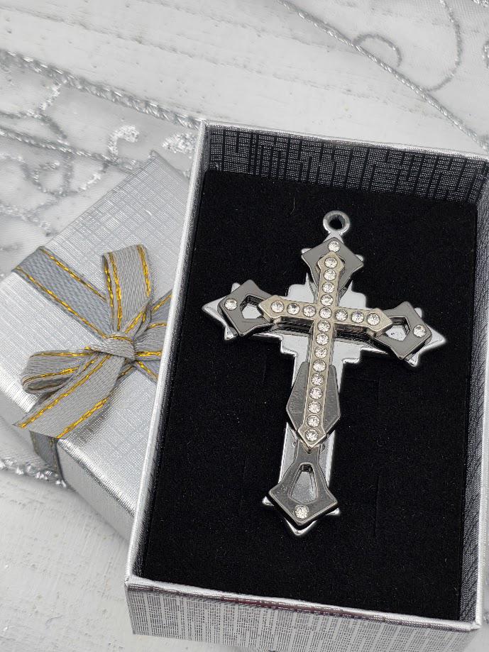 A silver cross pendant with rhinestones is displayed in a silver box with a gold ribbon bow.