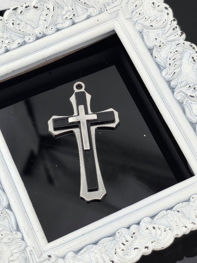 A silver cross pendant is displayed in a white ornate frame against a black background.