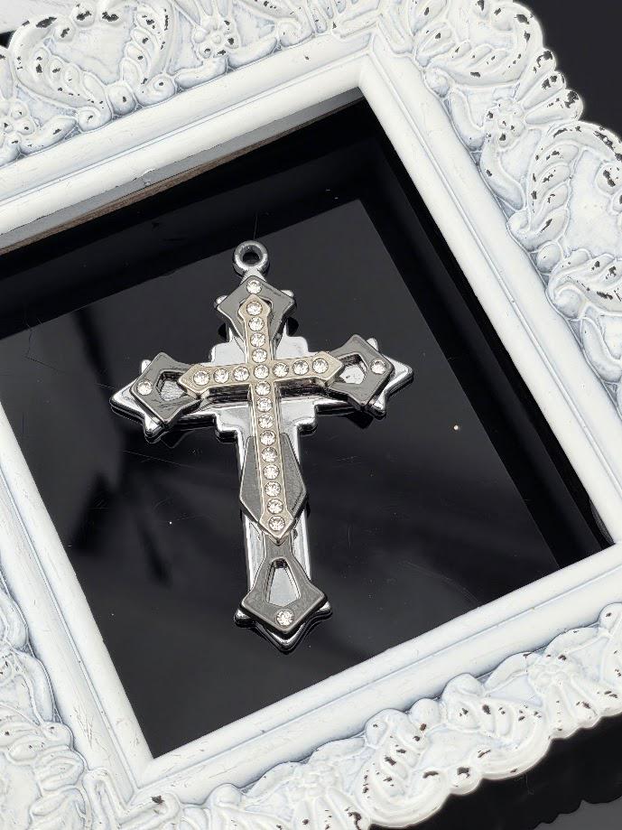 A silver cross pendant with rhinestones is displayed in a white ornate frame against a black background.