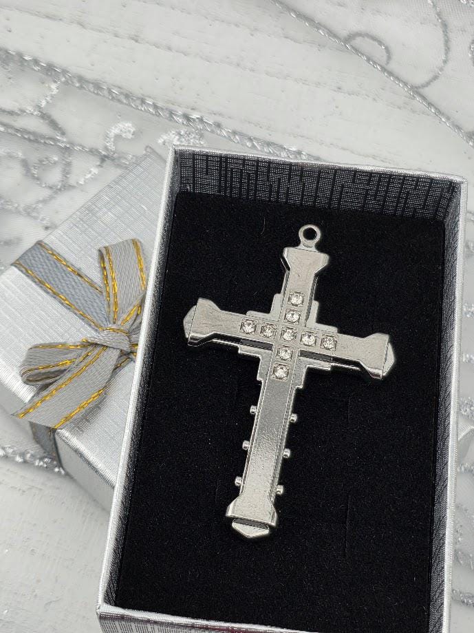 A silver cross pendant with small rhinestones is displayed in a silver box with a gold ribbon bow.