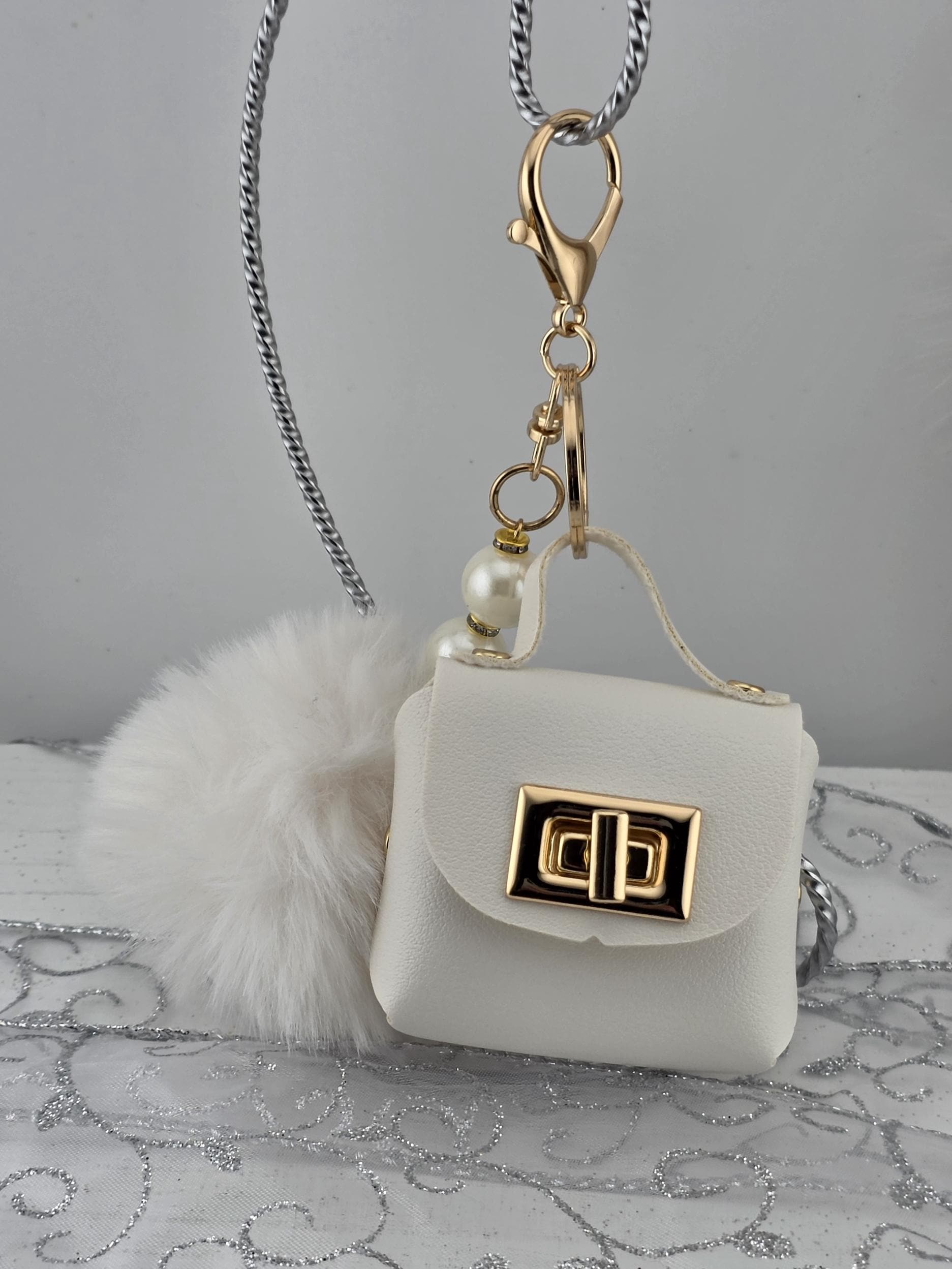 A white leather handbag with a gold lock and a fluffy white pom pom charm attached to it.
