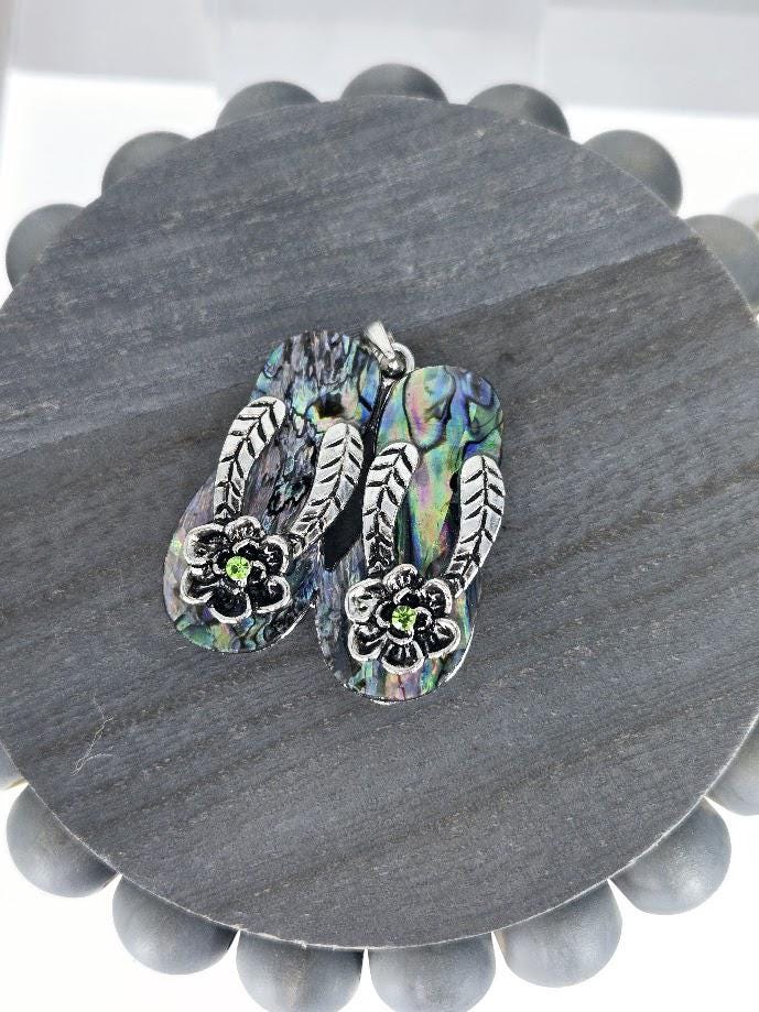 A pair of silver-toned flip-flop-shaped earrings with colorful floral designs on the soles, placed on a gray circular surface.