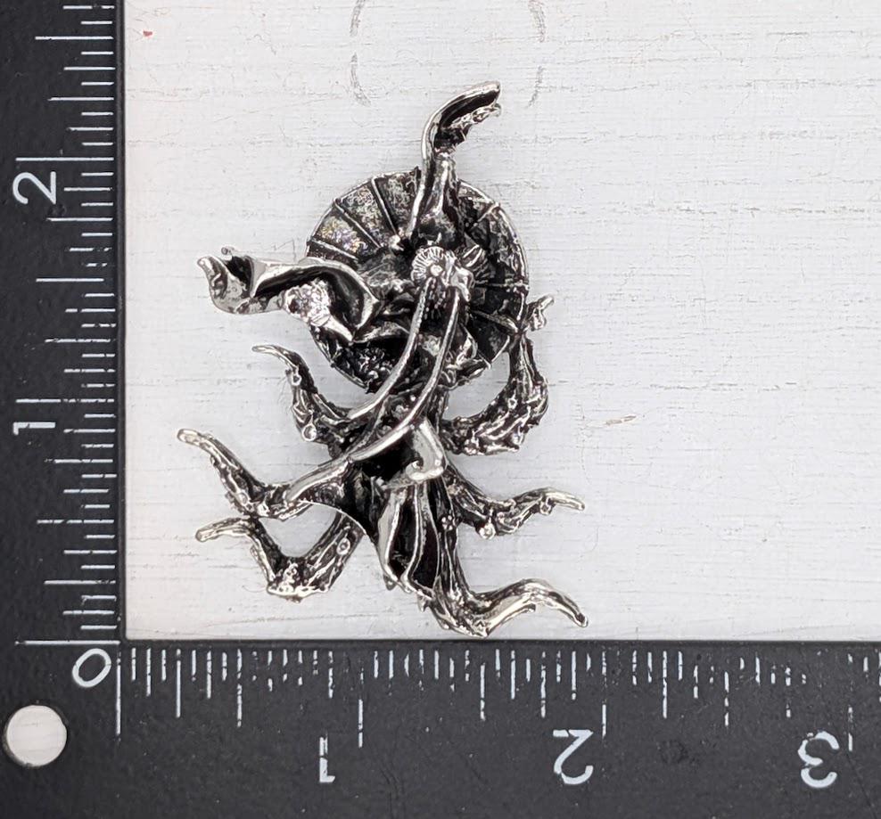 a silver-colored metal sculpture or brooch with an intricate design, placed on a ruler for size reference.