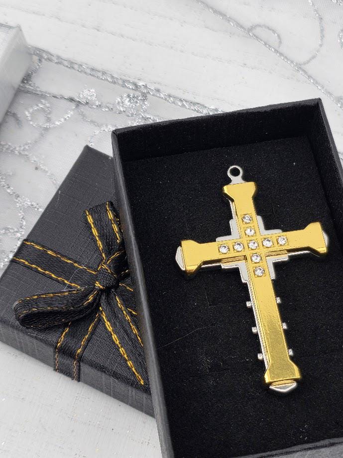 A gold cross pendant with rhinestones is displayed in a black box with a gold ribbon bow.