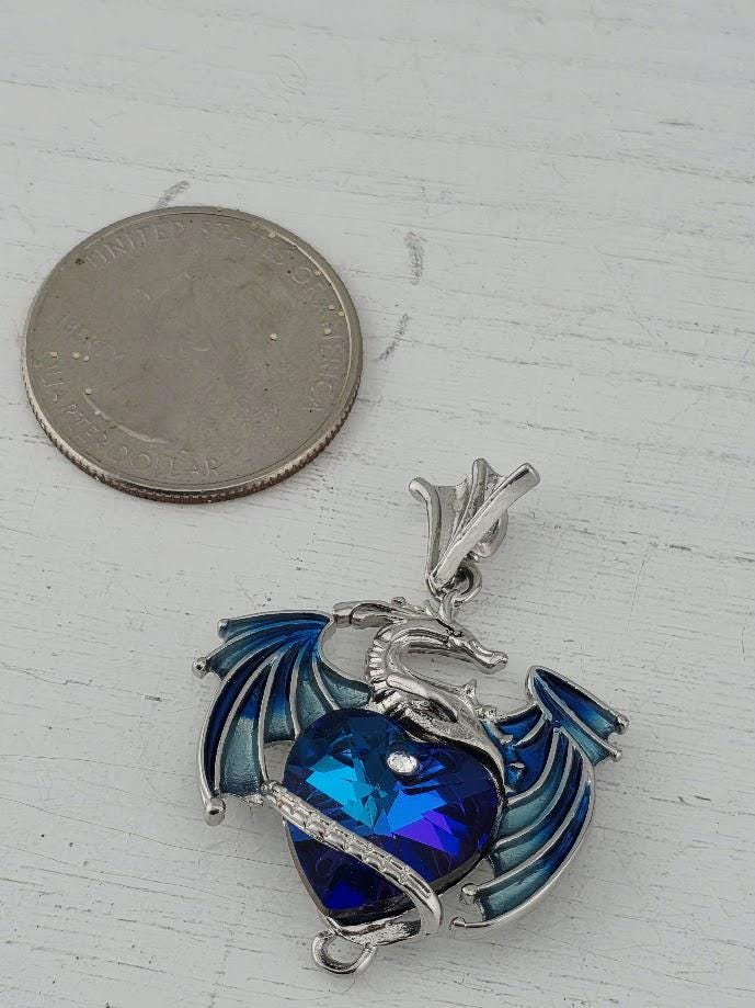 A pendant in the shape of a blue dragon with a heart-shaped gemstone is shown next to a United States quarter coin.