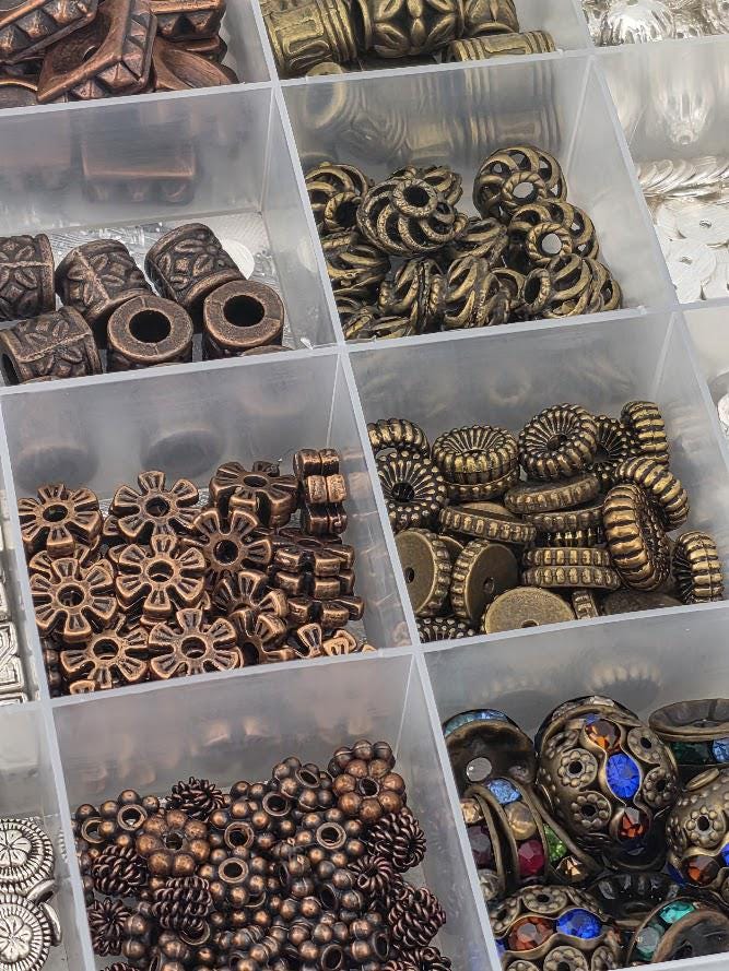 A collection of various beads and metal pieces, including copper-colored beads, gold-colored beads, and other decorative elements, arranged in multiple transparent containers.