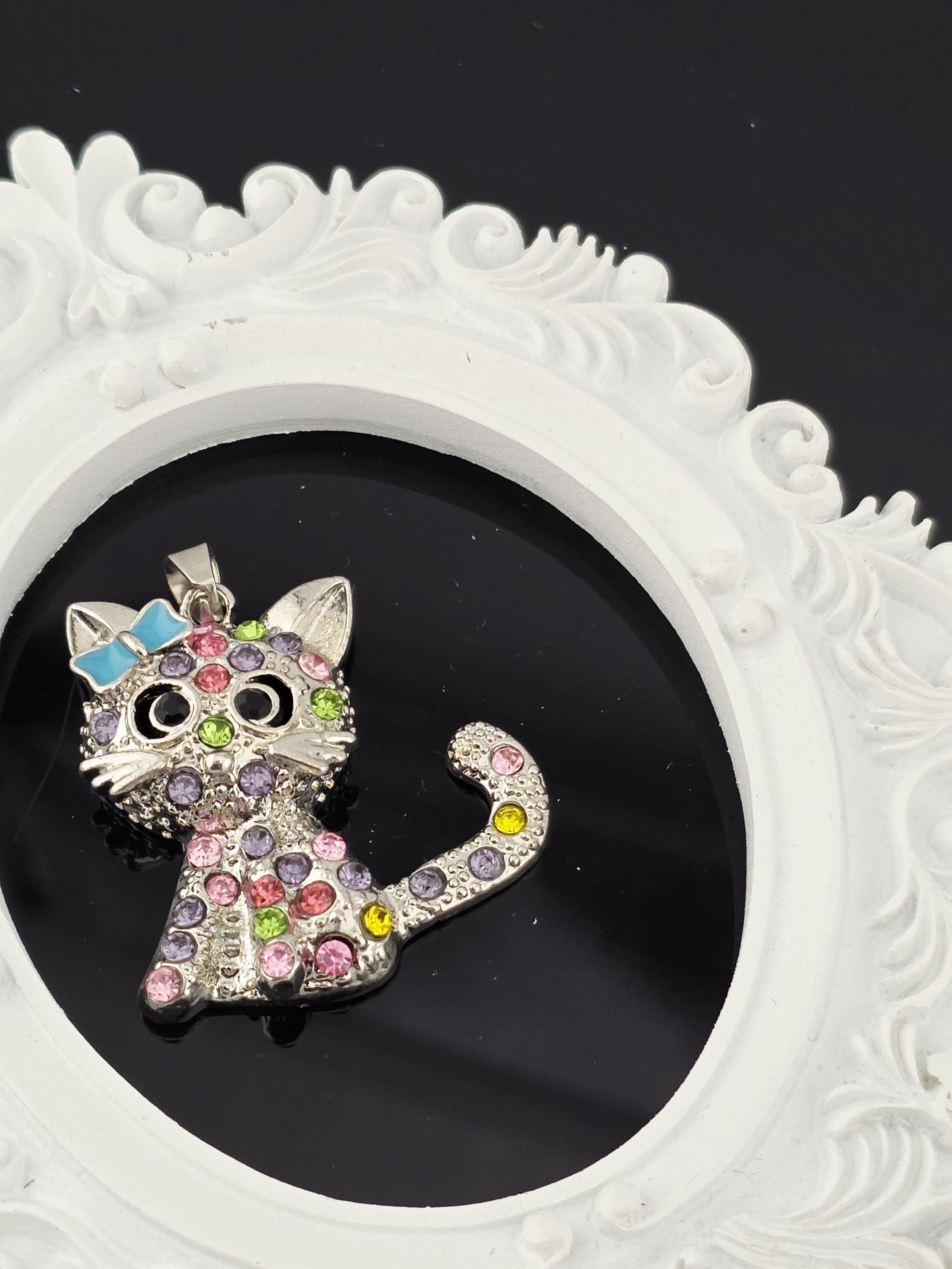 A decorative cat-shaped brooch with colorful gemstones is displayed against a black background, framed by an ornate white border.