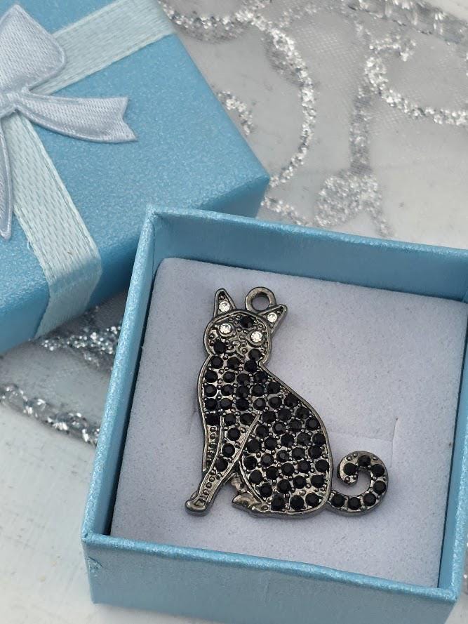 A silver-colored pendant in the shape of a cat, adorned with black gemstones, is displayed in a red gift box.