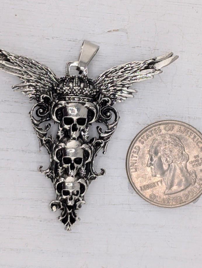 A silver pendant with three skull designs and wings, displayed in a black box with a white ribbon.