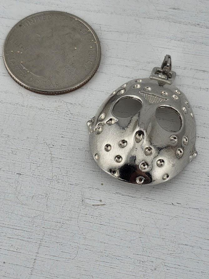 A silver-colored pendant in the shape of a mask is placed next to a United States quarter coin on a white surface.
