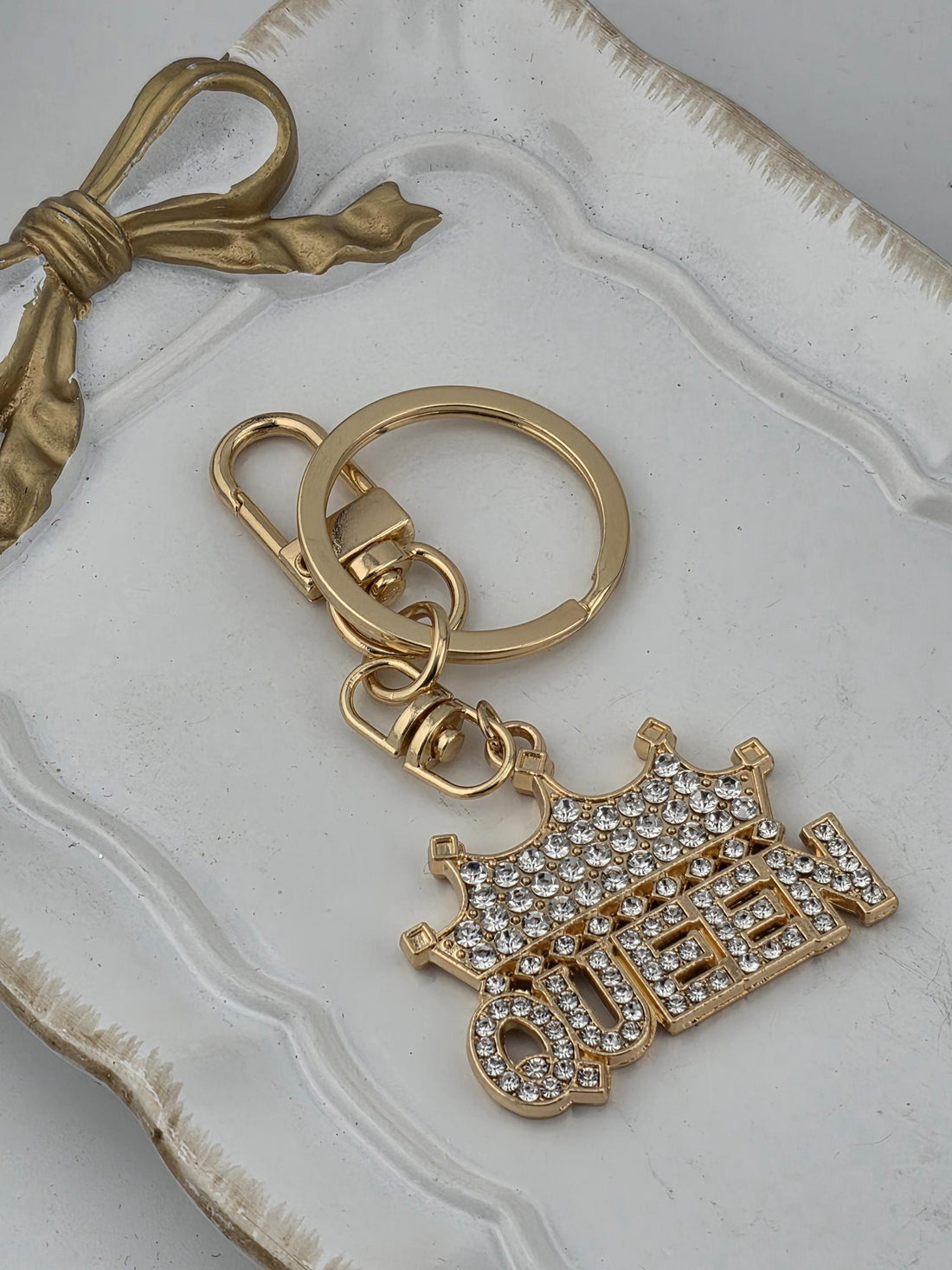 A gold-colored keychain with a charm that says "QUEEN" in a decorative font, made of small rhinestones or crystals.