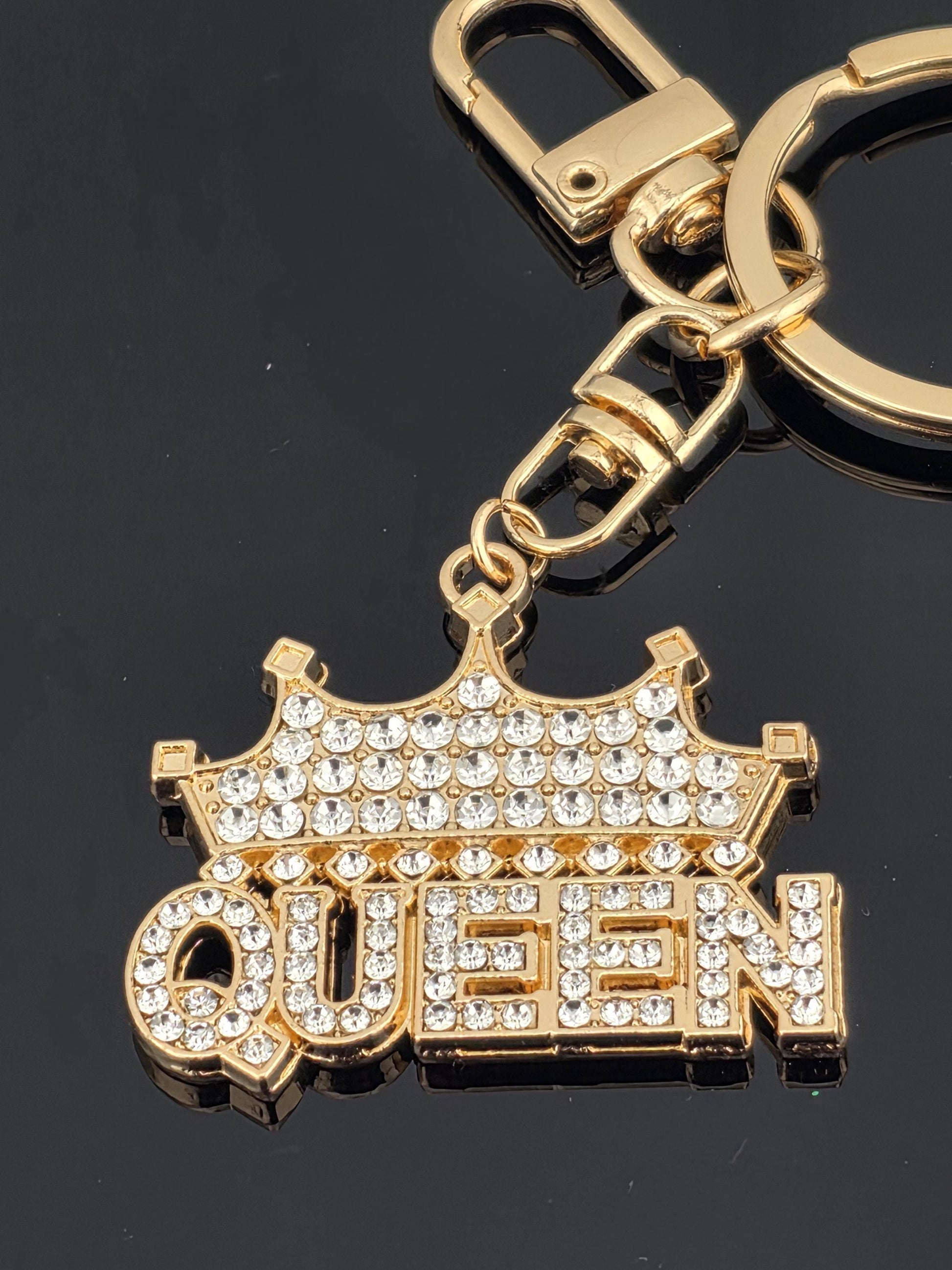 A gold-colored keychain with a large, sparkling rhinestone-encrusted design that says "QUEEN" on it.
