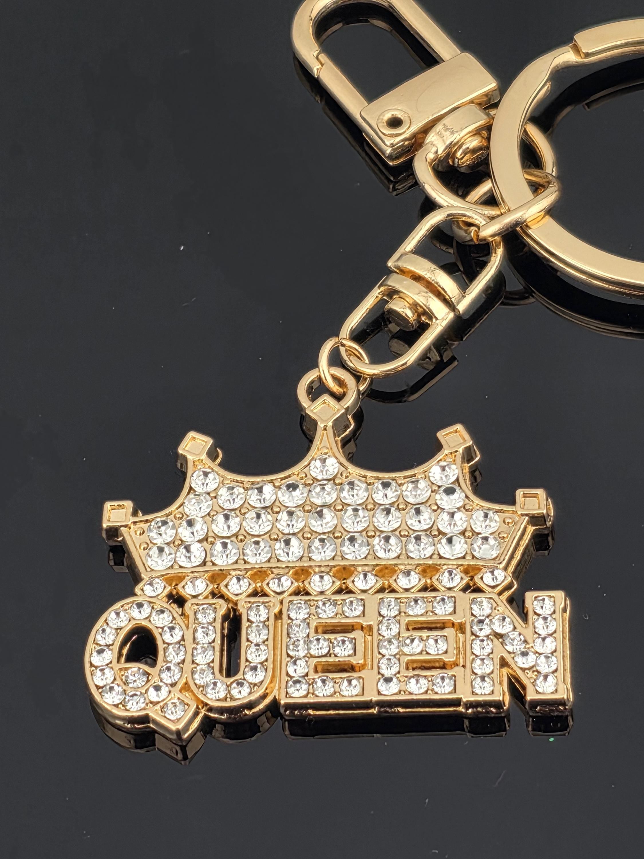 A gold-colored keychain with a large, sparkling rhinestone-encrusted design that says "QUEEN" on it.