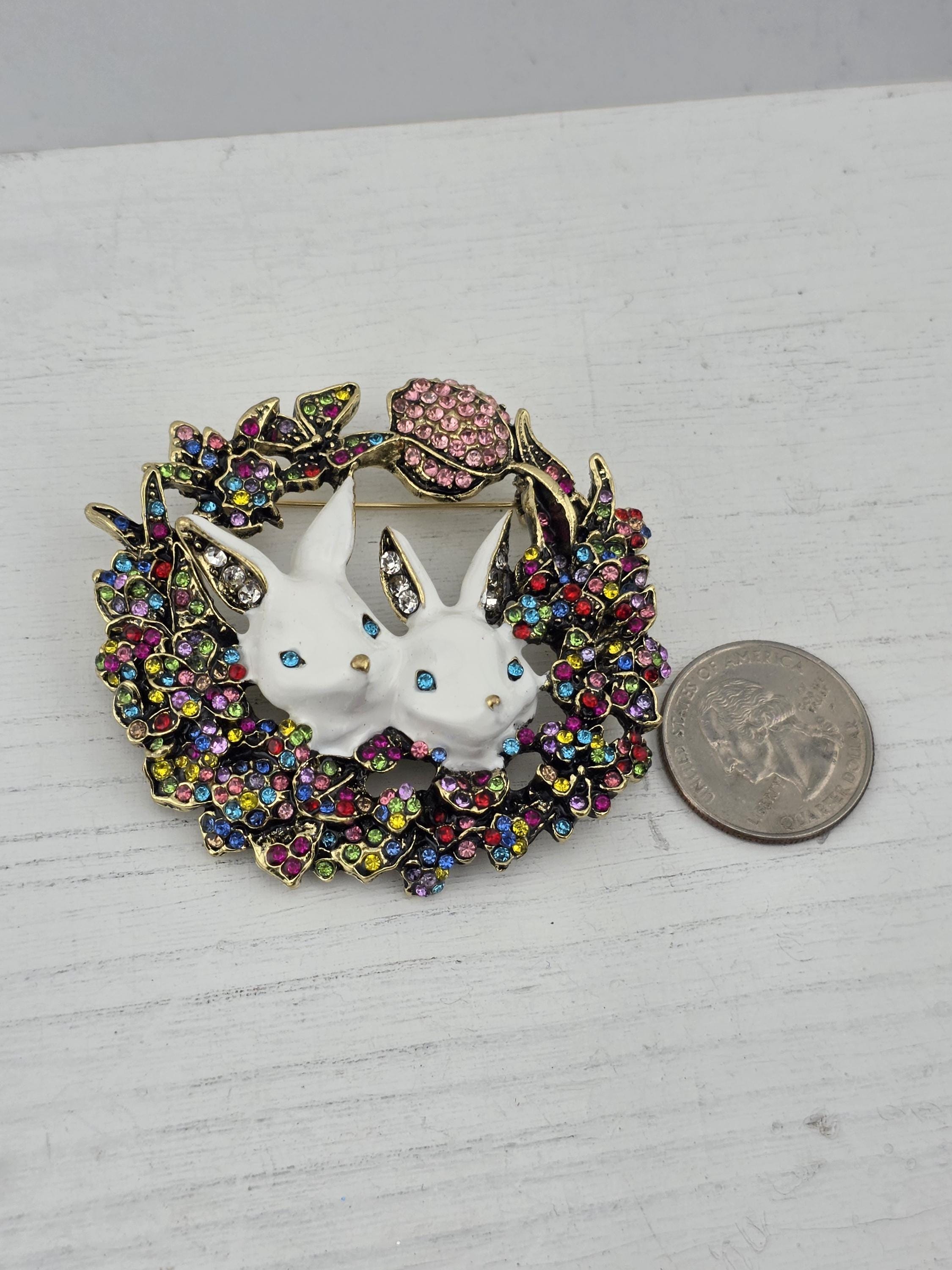 A colorful, ornate brooch with a white rabbit design and a wreath of flowers and leaves, placed on a white surface next to a coin.