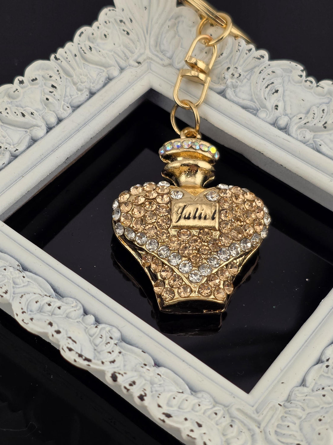 A decorative gold-colored perfume bottle with a heart-shaped design and sparkling rhinestones is displayed in a white ornate frame.