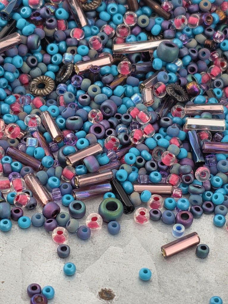 The image is a colorful assortment of beads and glass pieces, including various shades of blue, purple, pink, and black, scattered across a surface.